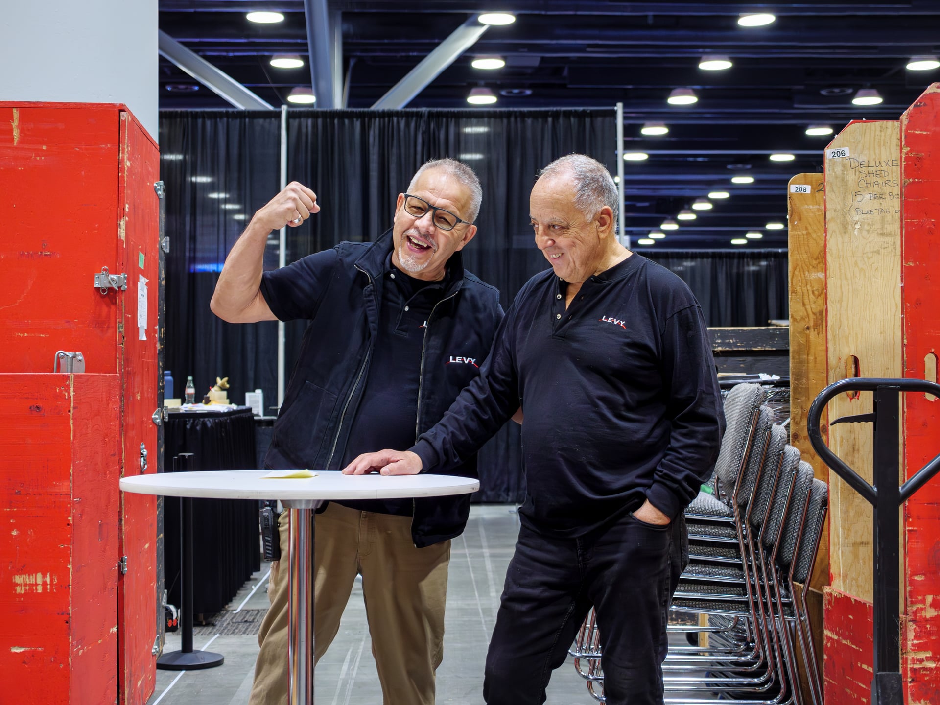 Two senior members of the LEVY team in branded polos sharing a laugh on the show floor at the end of the day, with stacked chairs and red equipment carts behind them.