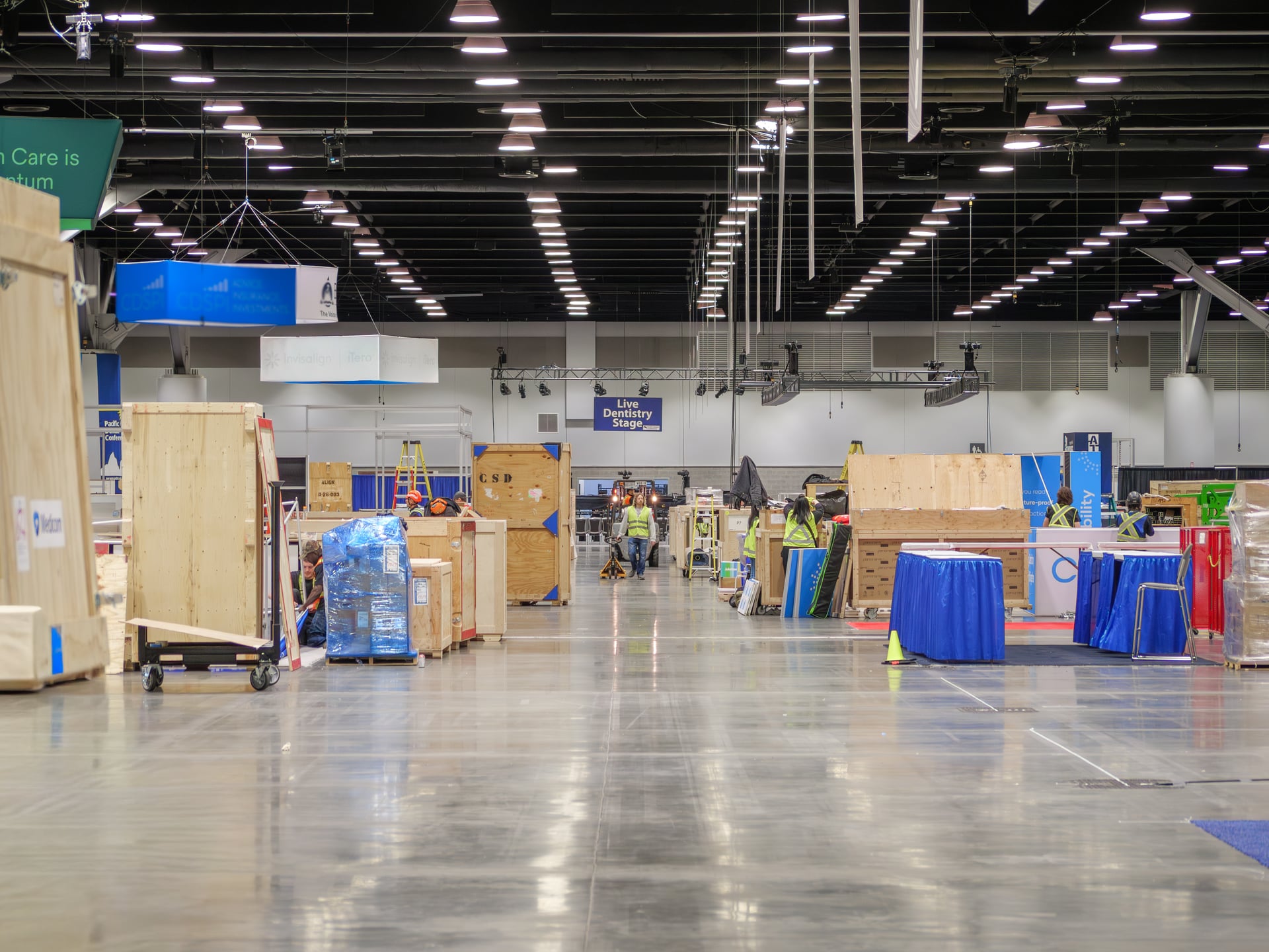 Wide aisle shot of the convention floor during build-out — wooden crates, blue drape kits, and a small crew in safety vests.