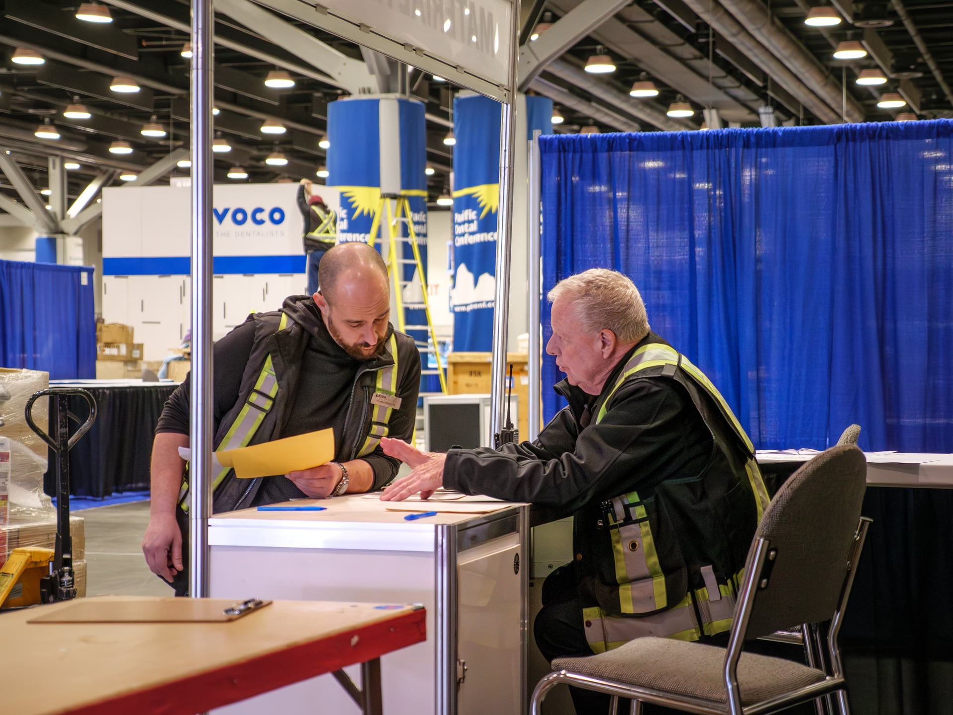 Two LEVY crew members in yellow safety vests reviewing show paperwork at a booth-in-progress, with VOCO signage visible.