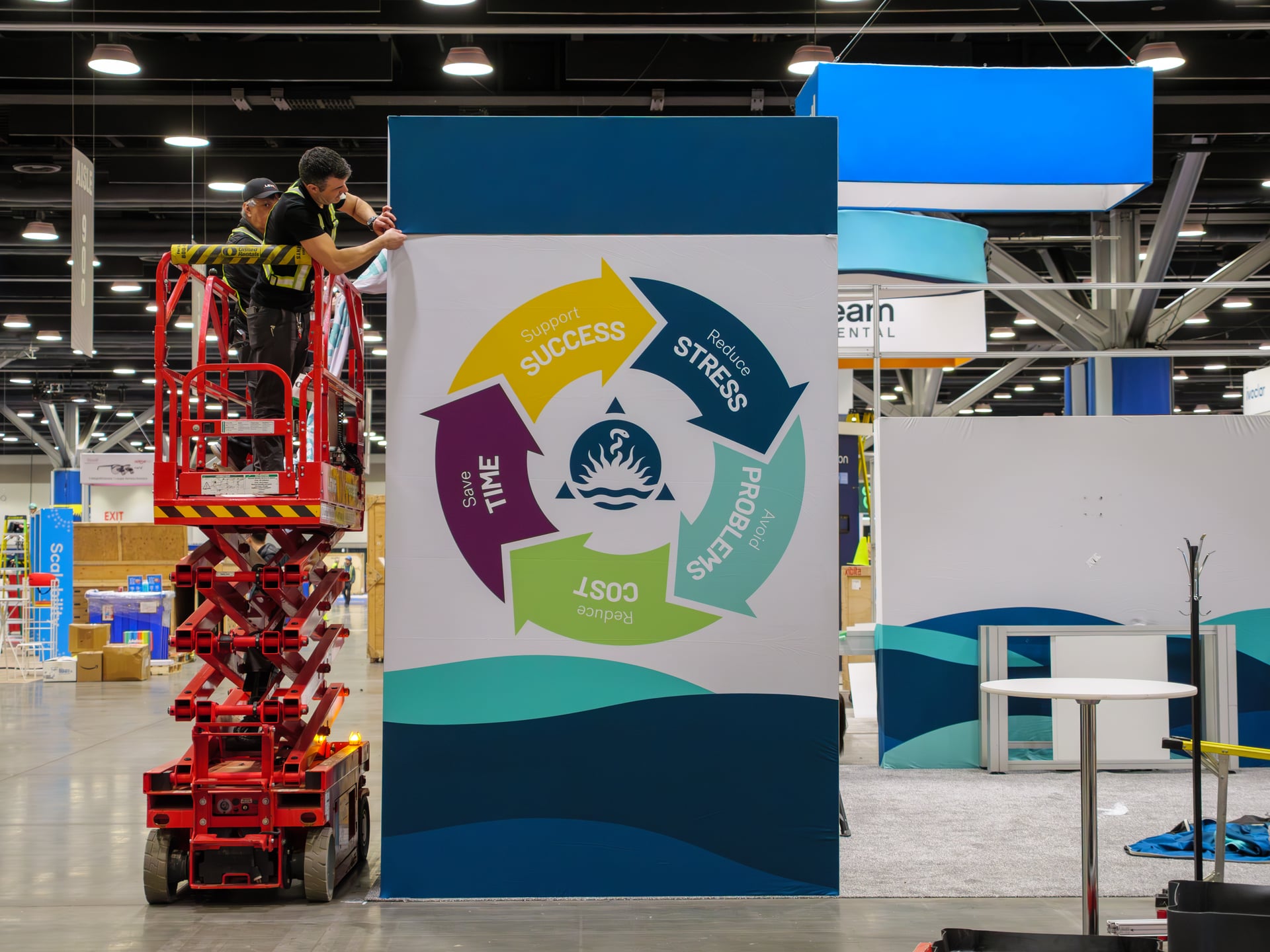 A LEVY crew member on a red scissor lift, installing a large branded graphic panel for an exhibitor.