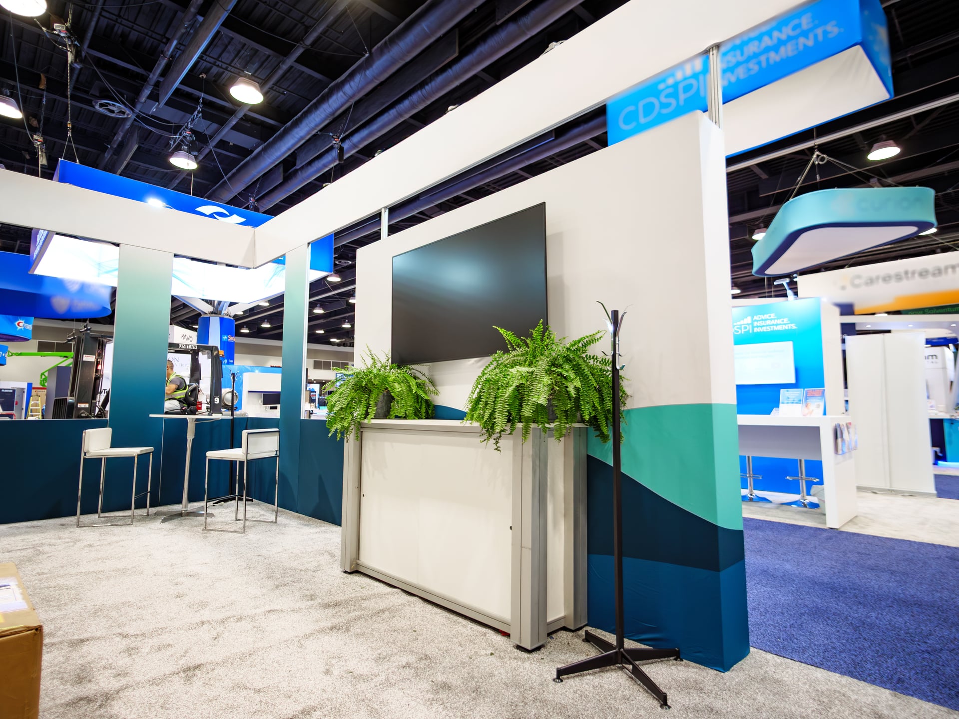 A finished CDS-Plus Insurance Investments booth, hardwall with a central screen, two ferns and bar-height seating.